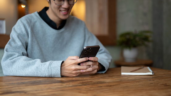 Cropped image of a happy young Asian man enjoys chatting with his friends or enjoys a new mobile application on his smartphone while relaxing in a coffee shop. People and technology concepts