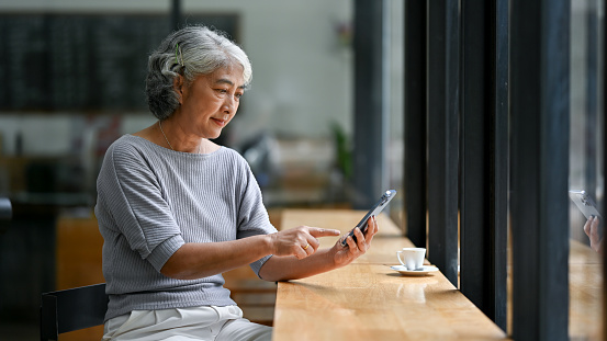 A relaxed and happy Asian retired lady in casual clothes enjoys the app or scrolls on social media on her smartphone while sitting in a coffee shop. Retirement lifestyle concept