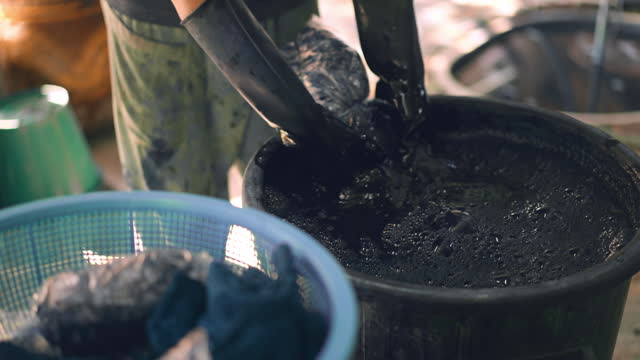 Local person's hands wearing black rubber gloves are dyeing a cloth with dark blue pigment in a jar containing pigment solution and using hands to squeeze the cloth for making dyeing process uniformity, traditional dyeing process in rural area