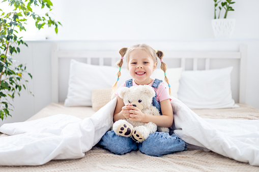 a small child, a blonde girl, holds a teddy bear in her hands under a blanket and smiles on a white bed in a bright bedroom.