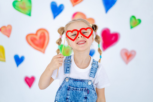a small child blonde girl with red glasses in the form of hearts smiles against the background of colorful hearts in the background, the concept of love and Valentine's day.