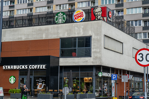Cafe store Starbucks coffee shop, KFC and Burger King under one roof. People are walking. Logos of competing companies. Street outdoor. Showcase sign. Poland, Warsaw - July 28, 2023