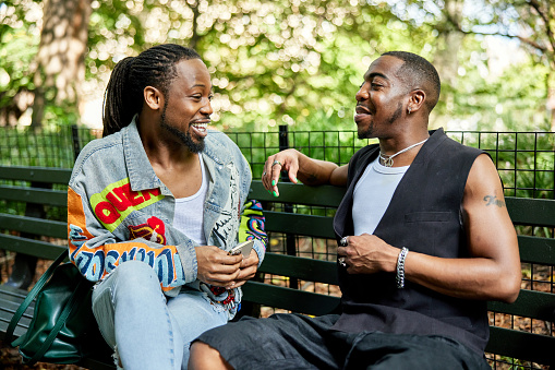 Gay Black New Yorkers in late 20s and early 40s sitting on bench, laughing, interacting face to face, summertime.