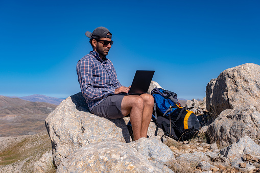 30-year-old model with a backpack. Wearing a hat and sunglasses. In a steep, mountainous area at high altitude. Open air. Mountain, nature view. Male model using a laptop. Getting his work done. Freelancer. Video calling is a multi-purpose topic.He has a hot water thermos and a backpack with him. Trekking at high altitudes. Exploration and adventure.