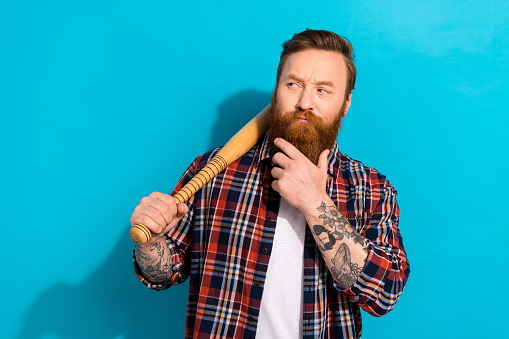 Photo of minded suspicious man hand on beard dressed checkered shirt look empty space bat on shoulder isolated on blue color background.