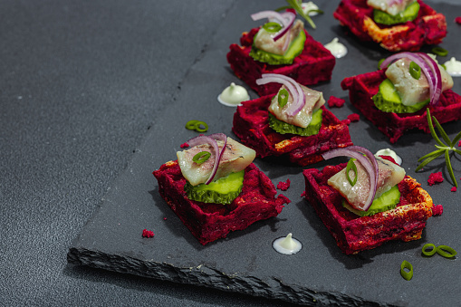 A delicious festive appetizer with herring on pieces of beet waffles with spices and sauce. Trendy hard light, dark shadow, black stone concrete background, close up