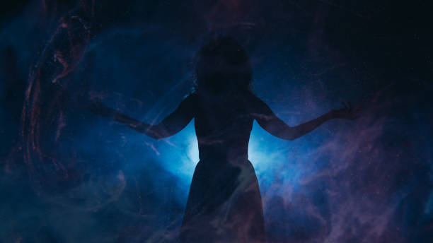 Spotlight silhouette. Spiritual energy. Woman with spread arms in haze shadow blue projector glowing particles fog dark background copy space.