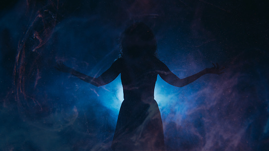 Spotlight silhouette. Spiritual energy. Woman with spread arms in haze shadow blue projector glowing particles fog dark background copy space.
