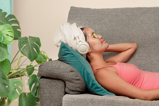 Side view of a woman lying on couch with cucumber slices under eyes and facial mask while relaxing during spa session at home