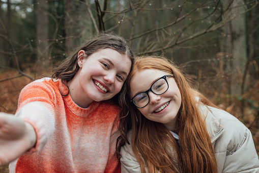 Close-up front view in Rothbury's woodlands, two female student teenagers are on an educational walk. They explore nature, blending lessons with the outdoors. They are using a smartphone to take a selfie.