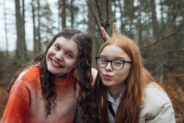Close-up front view in Rothbury's woodlands, two female student teenagers are on an educational walk. They explore nature, blending lessons with the outdoors. They are using a smartphone to take a selfie.
