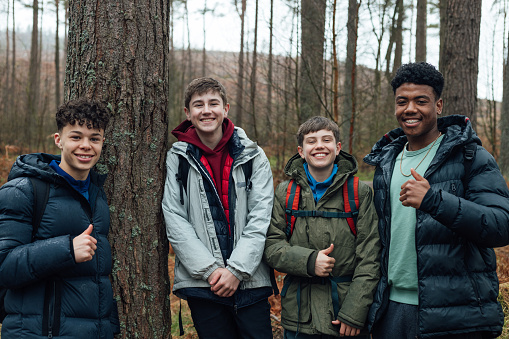 Front view three quarter length in Rothbury's woodlands, a group of male student teenagers are on an educational walk. They explore nature, blending lessons with the outdoors. They are smiling and looking at the camera.