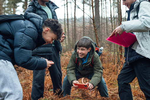In Rothbury's woodlands, a group of male student teenagers are on an educational walk. They explore nature, blending lessons with the outdoors. They are standing in a circle smiling while using a smartphone to take pictures and inspect the forest floor.