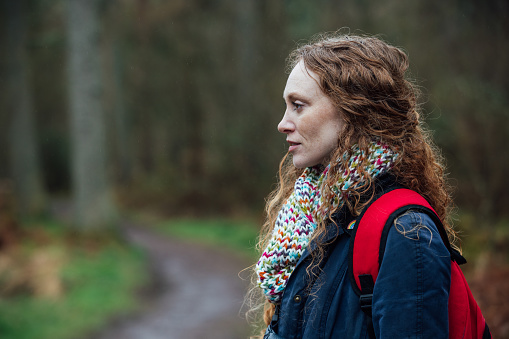 A waist-up side view in Rothbury's woodlands, a mid adult redhead woman is standing and looking into the distance, she is wearing warm clothing.