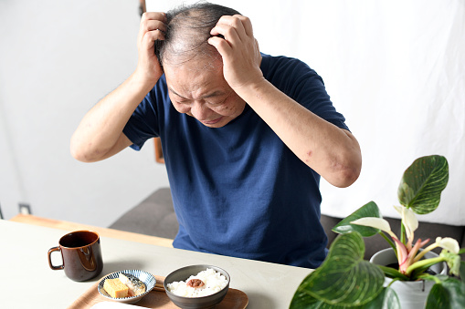 An elderly Asian man with a troubled face holding his head in front of a meal indoors