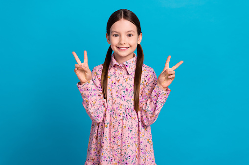 Photo of cute young girl show double v sign everyone unknown person wear dress first day kindergarten isolated on blue color background.
