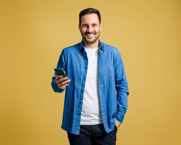 istock Smiling handsome young man browsing social media on mobile phone while standing on yellow background 1693477557