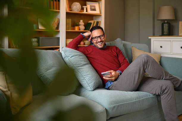 Handsome young entrepreneur smiling and scrolling smart phone while relaxing on sofa in living room