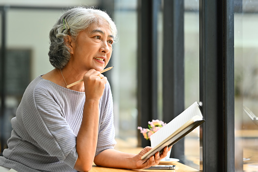 Beautiful gray haired mature woman holding book and looking away, daydreaming or thinking on good retirement plans.