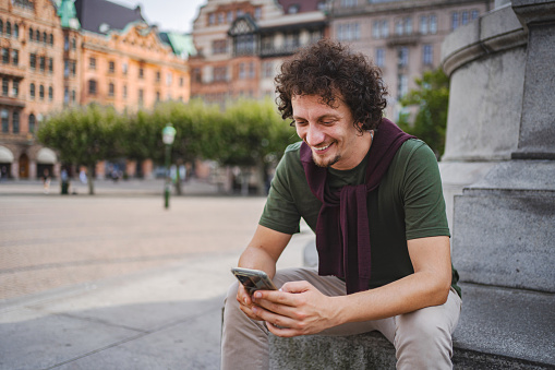 Men with curly hair looking at his phone while relaxing on the steps in the city square