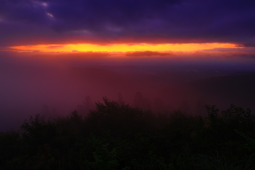 Stunning sunset with sky filled with clouds is beautifully painted with hues of orange reflecting the setting sun, trees provide a contrasting silhouette against the fiery sky