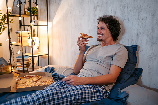 Handsome young man eating pizza in the bed at night in his bedroom.