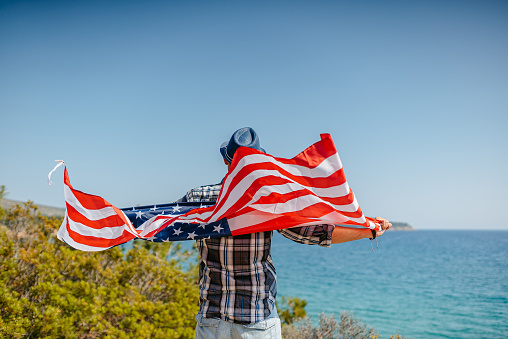 Young man holding a USA flag while looking at view of the coastline in Thasos, Greece.