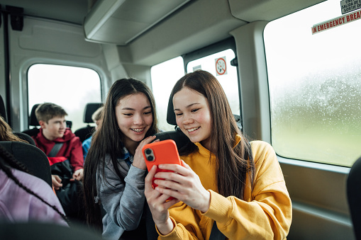 A front view of a two teenage girls on the minibus on the way to go on a hiking field trip. They are looking at one of the girls mobile phones and smiling.