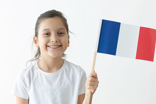 Little girl is holding French flag in hand and is looking at camera in front of white background.