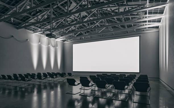 Side view of an empty darkened large gallery/showroom interior with empty seats (no people), and a wide projection screen in the middle for events under a ceiling industrial construction, illuminated by side wall reflectors