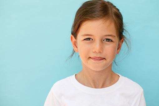 Little girl wearing white t-shirt is smiling and looking  at camera in front of blue\n background.