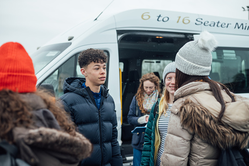 An over the shoulder view of a group of teenage high school students who are waiting around for the last of the classmates by the minibus before they head off for a school field trip to Rothbury in Northumberland in the North East of England.