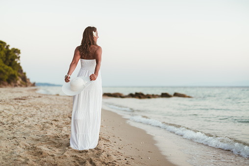 Rear view of a beautiful young smiling woman in white dress walking on the beach and holding white summer hat.