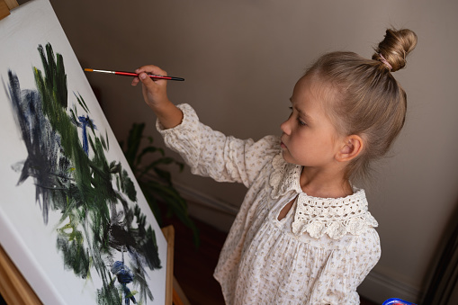 little cute girl draws with paints on canvas at the easel.