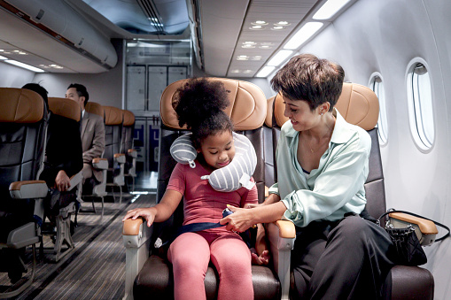 Parent take care kid on airplane, mother teach daughter to use seat belt during sitting in seat inside aircraft, African girl child with curly hair passenger traveling by plane, airline transportation