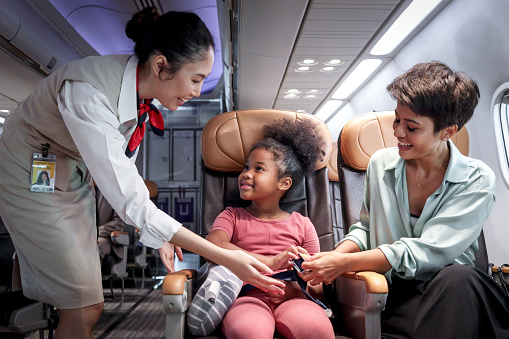 Friendly air hostess take care kid passenger on airplane, flight attendant teach curly hair African girl to use seat belt during sit in aircraft seat, child traveling by plane, airline transportation