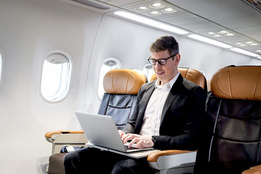 Busy businessman working on laptop computer during sitting in comfortable seat inside airplane, male passenger working in aircraft cabin during business trip, traveling with airline transportation.