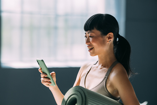 Getting ready for yoga class, an Asian woman holds her mat and checks her phone.