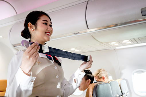 beautiful Asian female cabin crew air hostess training safety measures instruction to passenger, flight attendant stand aisle inside aircraft, holding seat belt to show safety rules before fly.