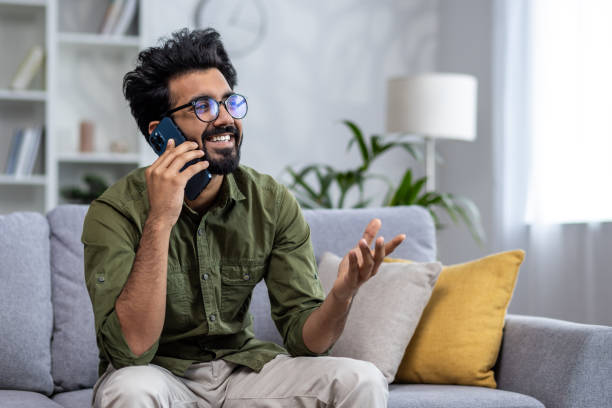 istock Young successful and smiling man talking on the phone while sitting on the sofa in the living room, Indian cheerfully chatting with colleagues and friends 1693760131