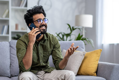 Young successful and smiling man talking on the phone while sitting on the sofa in the living room, Indian cheerfully chatting with colleagues and friends.