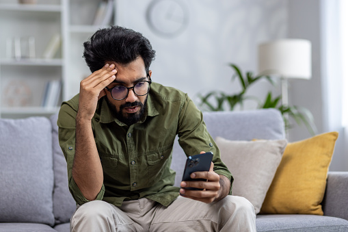 Upset and sad man sitting on sofa at home in living room, hindu man reading bad news online from phone, depressed and sad man