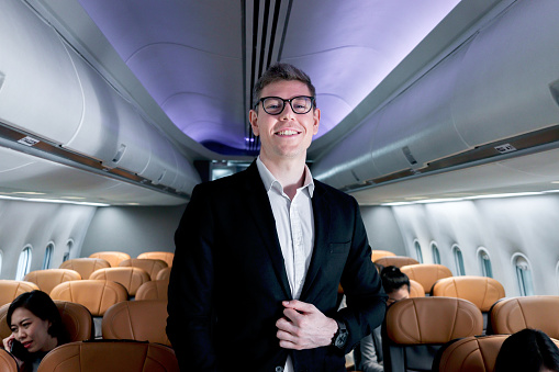 Portrait of happy smiling businessman in black suit, standing on aisle inside airplane, male passenger traveling on business trip by aircraft, businesspeople traveling with airline transportation.