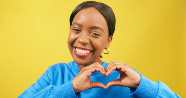 istock Young Black woman forms heart with hands, smiling yellow studio background 1693799758