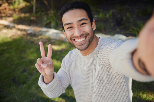In a picturesque park, a handsome man captures a selfie with his smartphone, and to add a playful touch, he raises his hand to form a peace sign. His smile radiates positivity, and the tranquil park serves as a beautiful background for this lighthearted moment of self-expression.