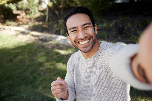 In a serene park setting, a handsome man smiles as he holds up his smartphone to take a selfie. The natural beauty of the park provides a picturesque backdrop for his photo, and his cheerful expression reflects the joy of capturing a memorable moment outdoors.