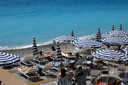 Nice, France - April 26, 2023: White-blue beach umbrellas and sunbeds set on a pebble beach for sunbathers to relax in both the shade and the sunlight here on the beach of the Promenade des Anglais.