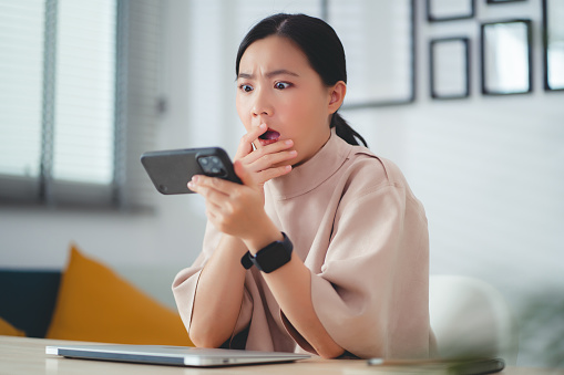 Asian woman feel scared and shock while watching something on smartphone sitting at home office.
