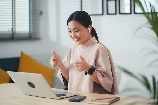 Asian woman wearing earbud showing thumbs up using laptop talking on video conference sitting  at home office. Happy woman working from home.