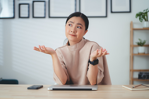 Asian woman thinking about project and feel confused, pointing to copy space sitting at home office.